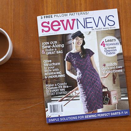 Sew News magazine on a wood surface beside a coffee cup, with a cover model in a pink plaid dress and sewing headlines