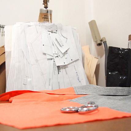 Sewing studio worktable with paper pattern pieces on a dress form, orange and gray fabric swatches, and metal buttons