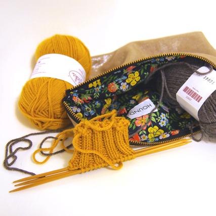 Knitting supplies: mustard yellow and gray yarn, wooden needles, in-progress sock, and a floral zippered project pouch