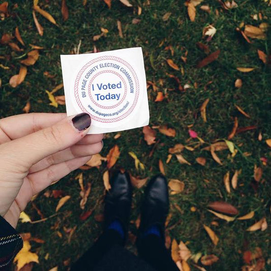 Hand holding I Voted Today sticker from DuPage County Election Commission above fall leaves and black boots on grass