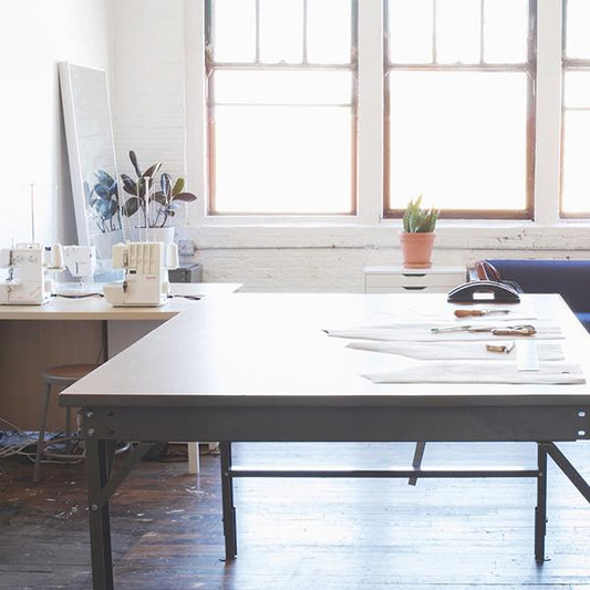 Grainline Studio workspace with large cutting table, pattern pieces, sewing machines, plants, and large industrial windows