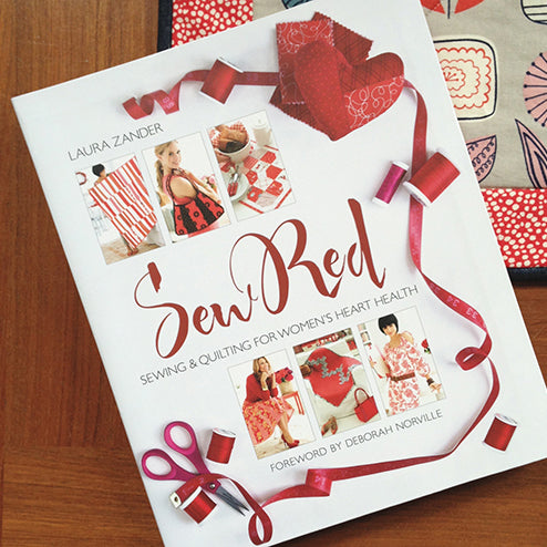 Sew Red book by Laura Zander on a wood surface, with red ribbon, pink scissors, thread spools, and fabric scraps
