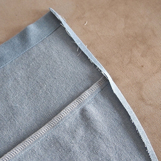 Close-up of a single folded hem on light-wash denim Alder Shirtdress fabric, showing a serged seam allowance and raw edge