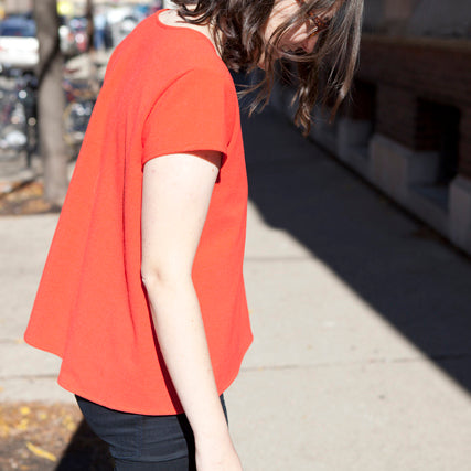 Person wearing coral-red Scout Tee with extended fuller back and short sleeves, paired with dark jeans