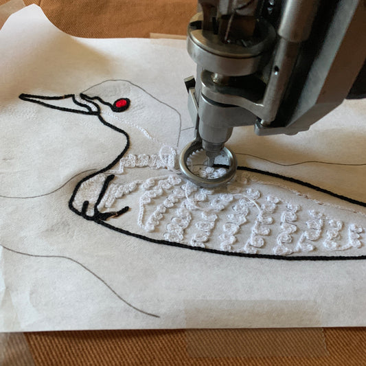 Embroidery machine stitching a loon on cream fabric with chainstitch, showing white body fill, black outline, and red eye