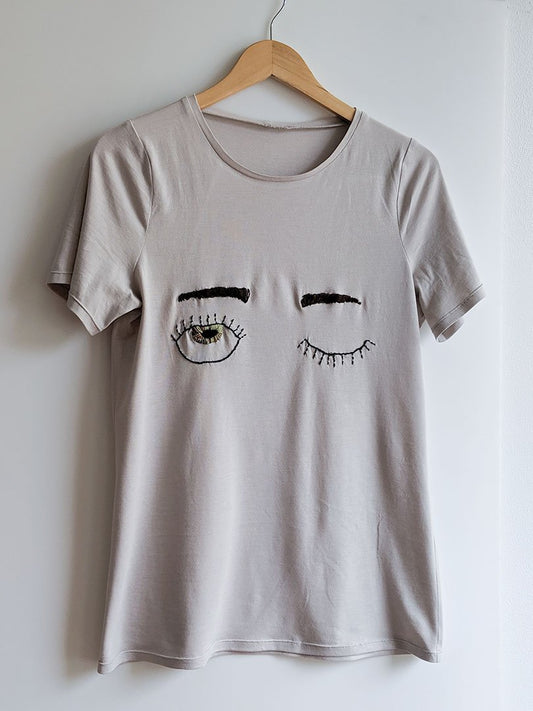 Light gray Lark Tee with hand-embroidered winking eyes design on wooden hanger with white background