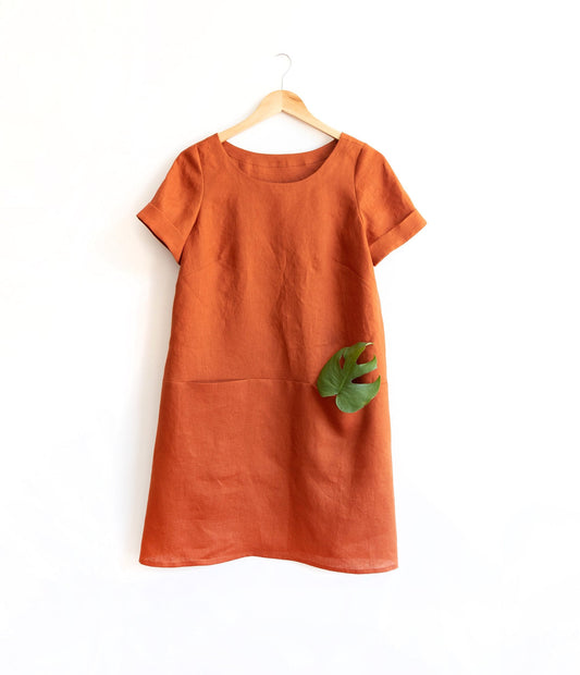 Uniform Tunic in rust linen with short cuffed sleeves and inseam pockets, monstera leaf tucked in one, on wooden hanger