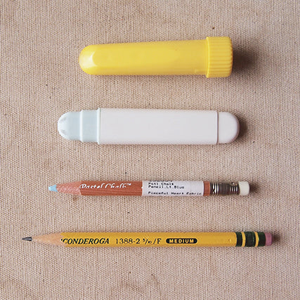 Four marking tools on beige fabric: yellow chalk wheel, white chalk wheel, blue chalk pencil, and yellow Ticonderoga #2 pencil