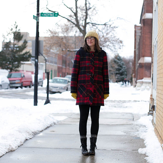 Person wearing red plaid Cascade Duffle Coat with black leggings, cream knit beanie, and black boots on a snowy urban street