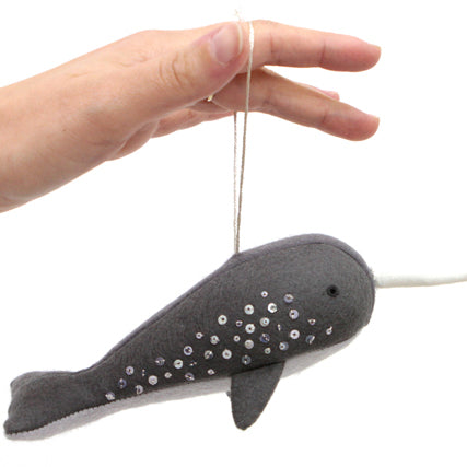 Hand holding a gray felt narwhal ornament with silver beaded details, long white tusk, and string loop for hanging