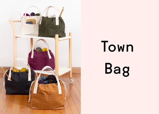 Five canvas Town project bags in natural, olive, plum, black, and toffee filled with yarn, styled on a wooden cart