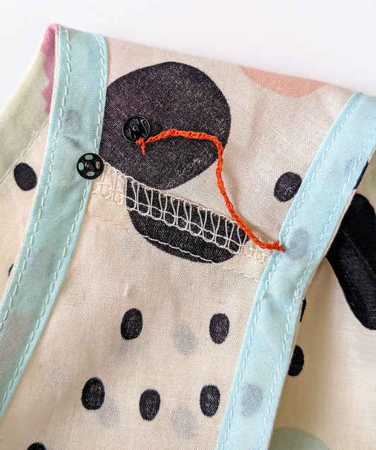 Needle pulling orange thread through fabric near armhole seam with serged edge and light blue binding.