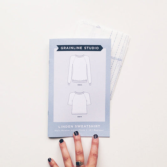 Grainline Studio Linden Sweatshirt pattern envelope showing two views with pattern pieces, hand with dark nail polish visible