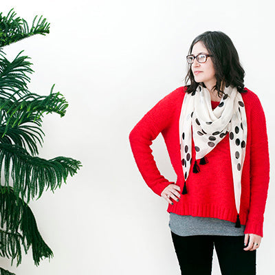 Woman wearing bright red sweater over gray top with polka dot tassel scarf, black pants, and glasses near a pine tree