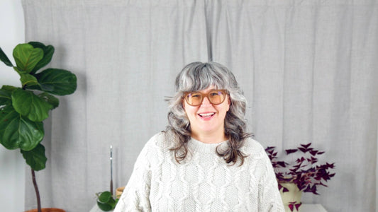Smiling woman with gray hair and glasses wearing a cream cable knit sweater surrounded by houseplants