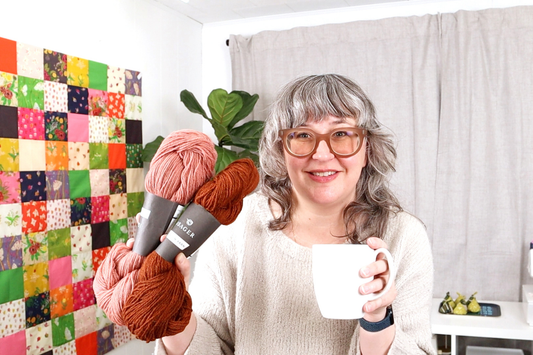 Woman in rose-colored glasses holding yarn and white mug with colorful patchwork quilt and plants in background
