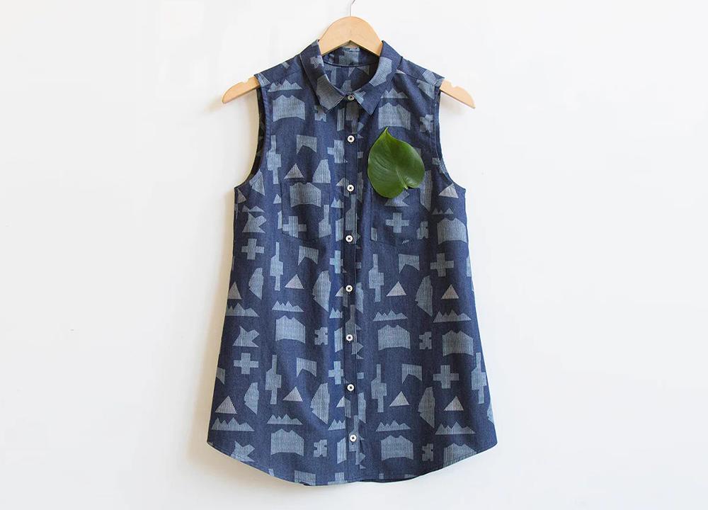 Alder tunic in blue geometric print denim hanging on wood hanger with green leaf in breast pocket showing shortened length
