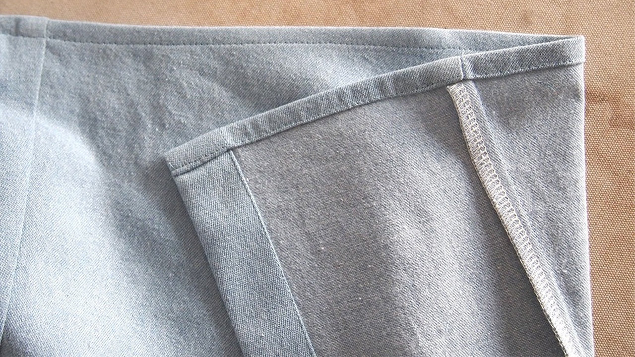 Alder hem on light blue denim showing double-fold technique with close edgestitching near inner fold on beige surface