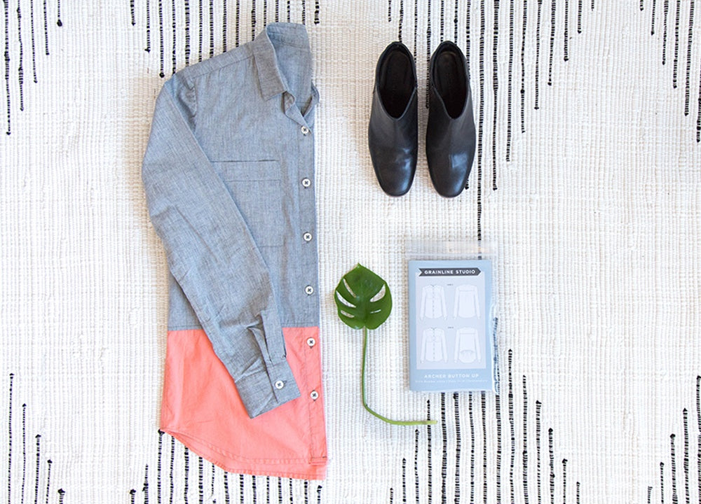 Gray Archer shirt with a coral color block at the hem laid flat beside black ankle boots, a monstera leaf, and the pattern