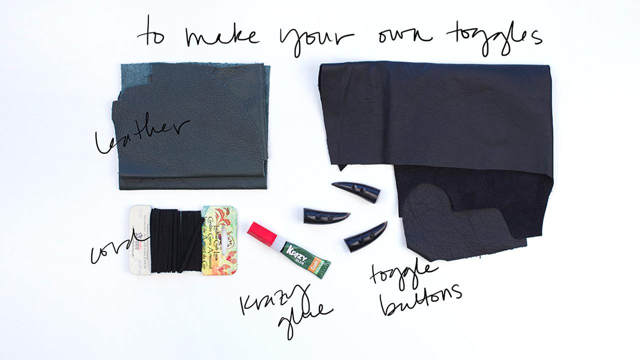 Toggle-making supplies: navy leather scraps, black leather cord, Krazy Glue tube, and three navy toggle buttons arranged on white surface
