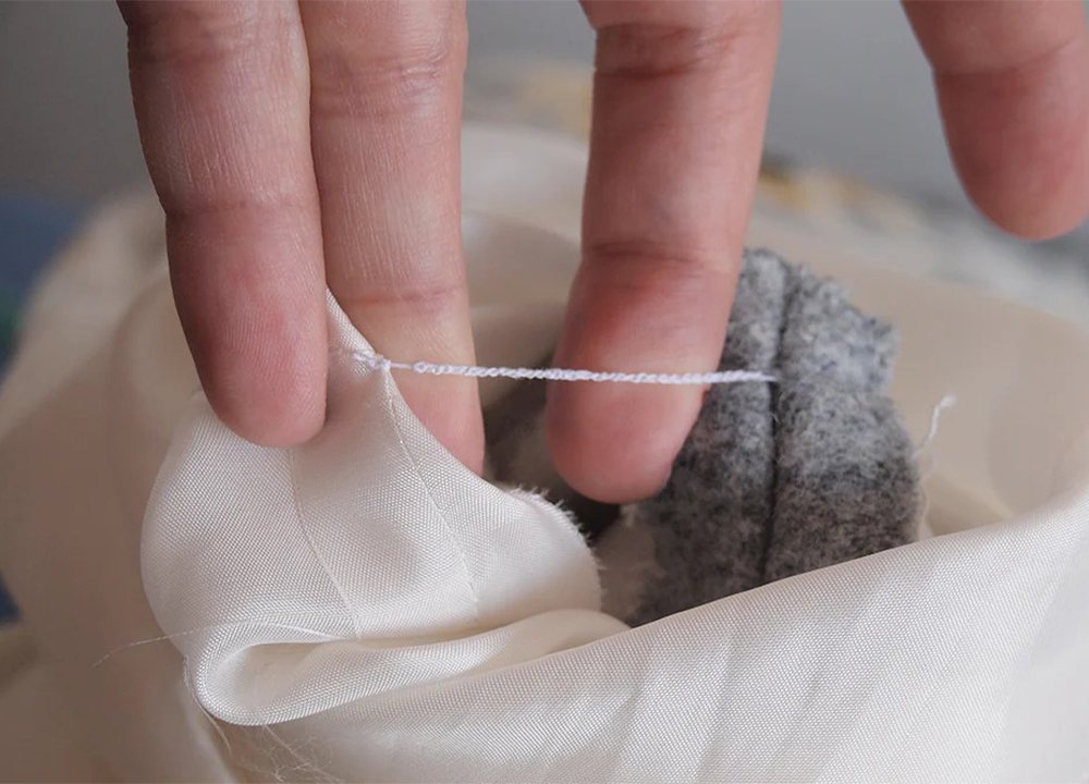 Hands demonstrating thread chain technique connecting white lining fabric to gray wool coat fabric at underarm seam