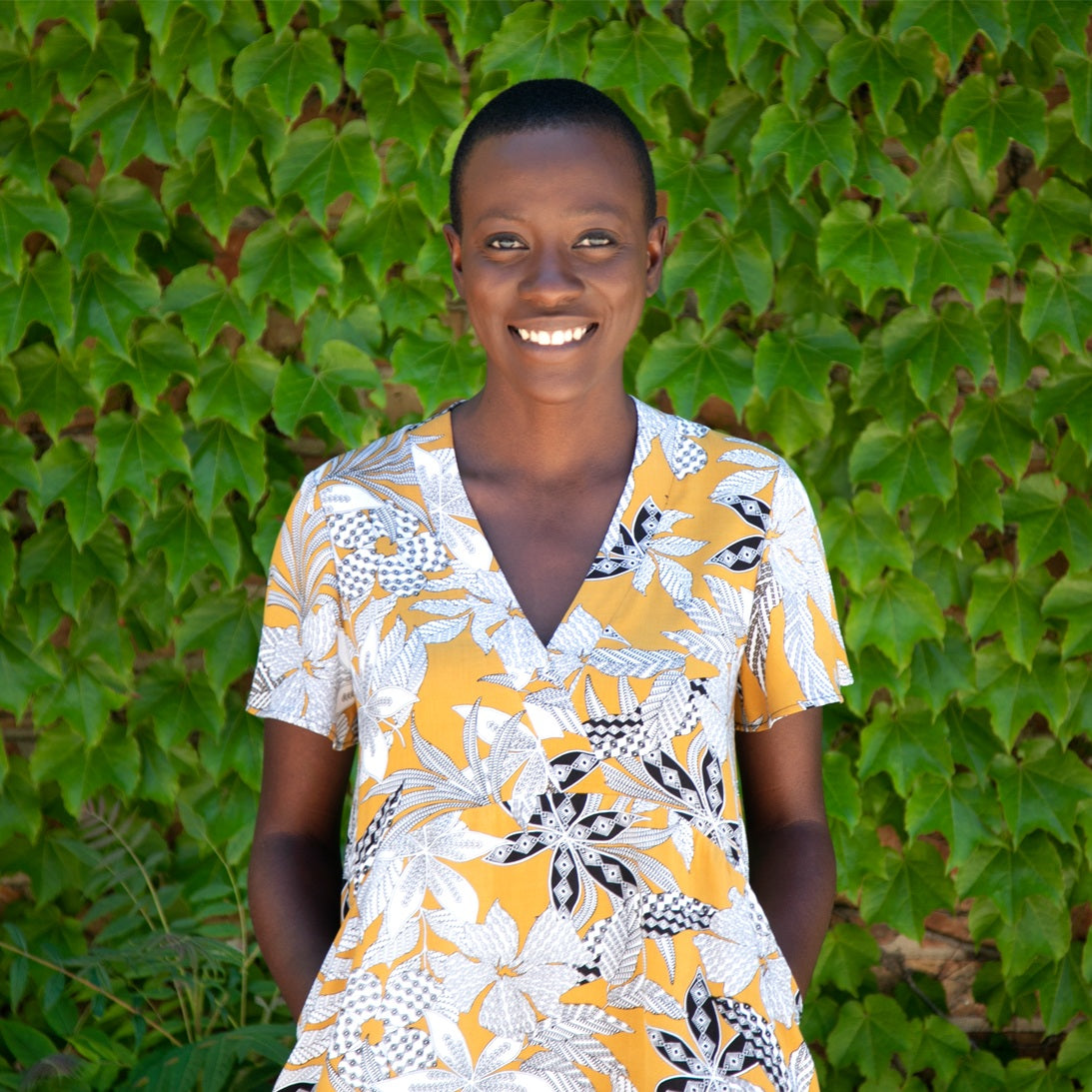 Model in Felix dress with V-neck, golden yellow fabric featuring black and white botanical print, ivy backdrop