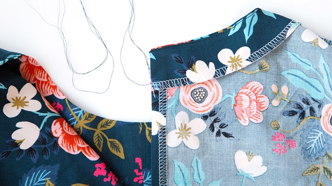 Hook and eye being hand-stitched to back neckline of completed Farrow Dress in navy and light blue floral rayon fabrics showing final closure attachment.