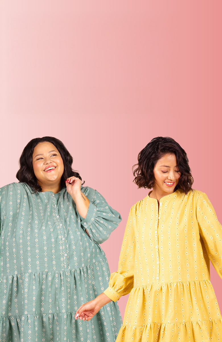 Two women in tiered dresses with floral print, one in sage green and one in yellow, smiling against pink backdrop