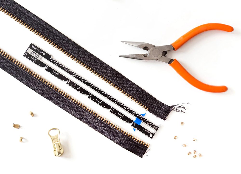 Zipper-making supplies: continuous chain with brass teeth, slider, stops, ruler, pliers, and removed teeth on white surface