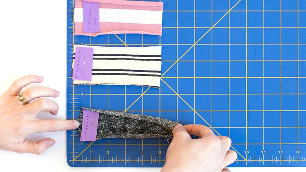 Hands stretching knit fabric swatches on cutting mat showing 20% stretch test: coral stripe, black stripe, and gray jersey
