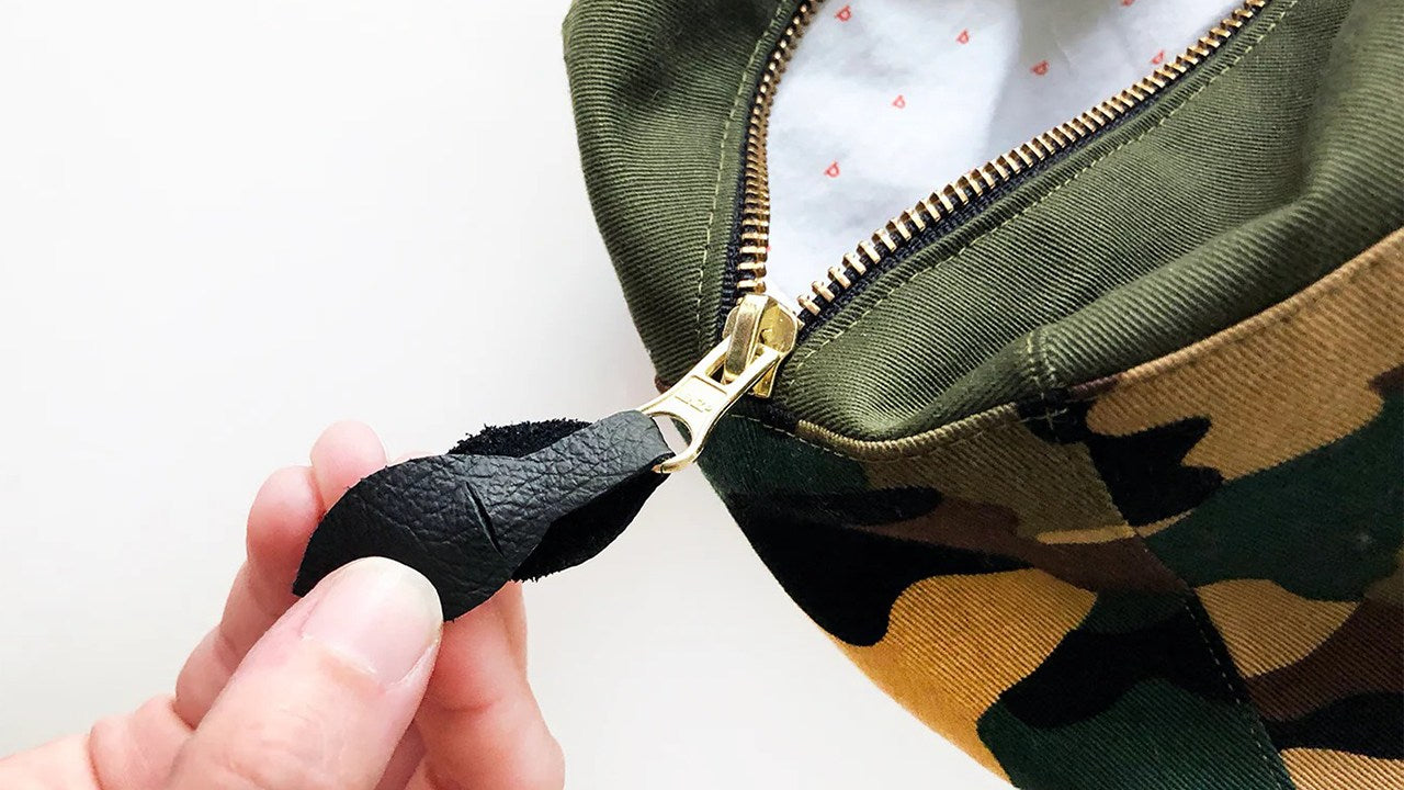 Hand holding black leather zipper pull attached to brass zipper on camo and olive green dopp kit