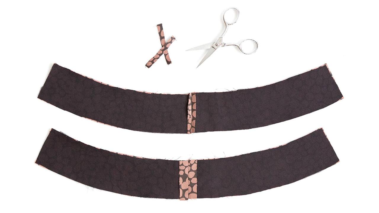 Two curved waistband pieces shown wrong side up with charcoal fusible interfacing and pink dot seam allowances, scissors above