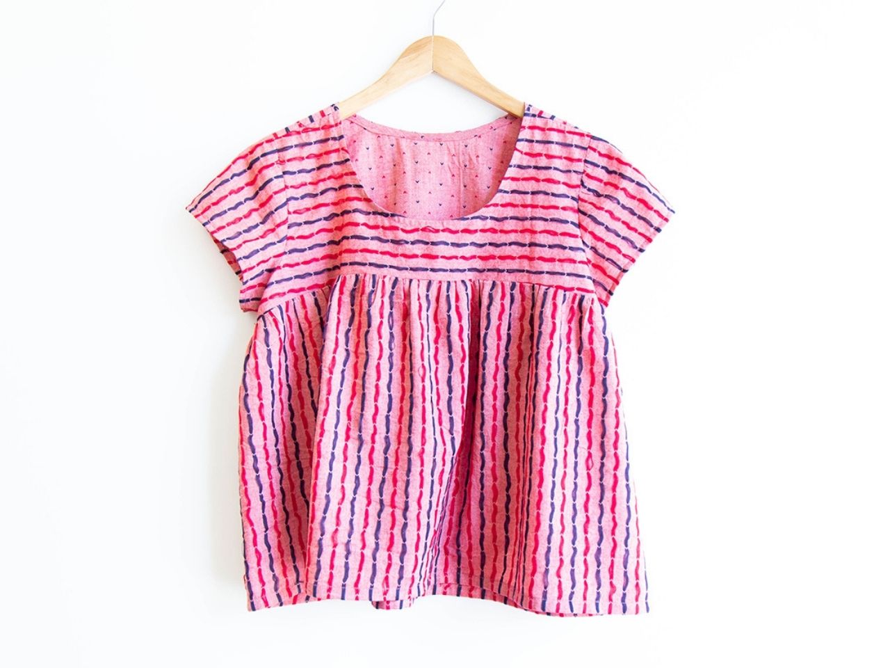 Scout Tee with gathered yoke modification in pink and red striped woven fabric with visible floats on a wooden hanger.