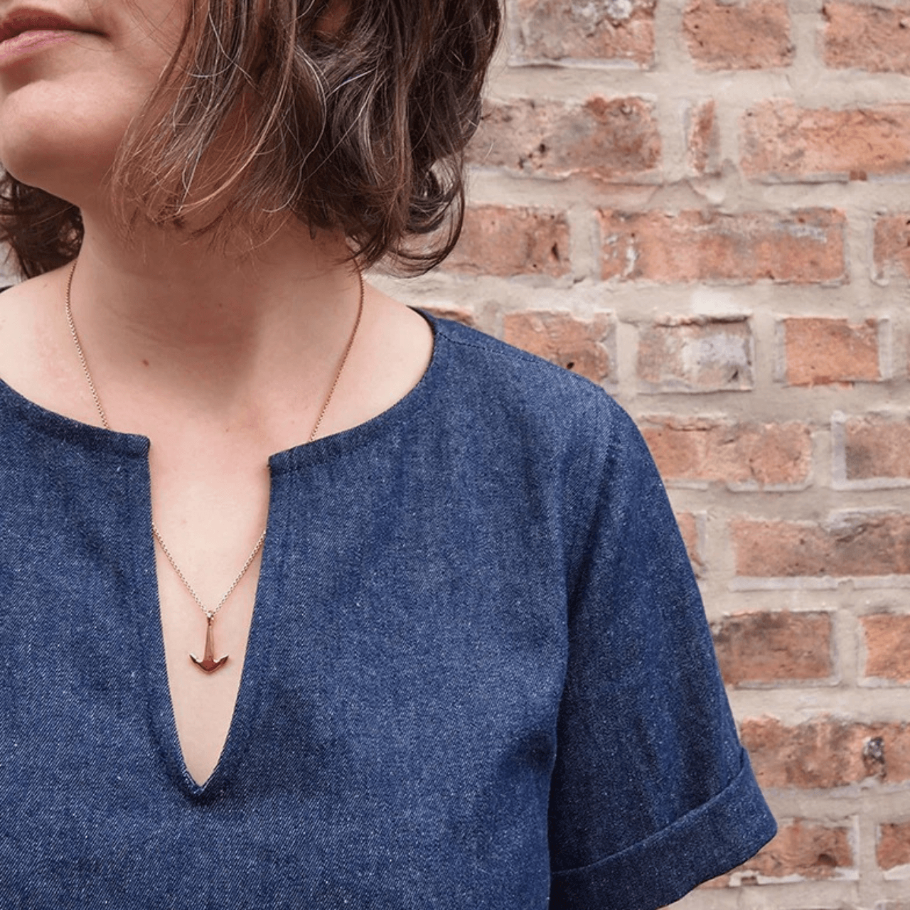 Close-up of a Scout Tee in denim with a V-neck opening and folded cuff sleeve, worn with a gold anchor necklace against a brick wall.