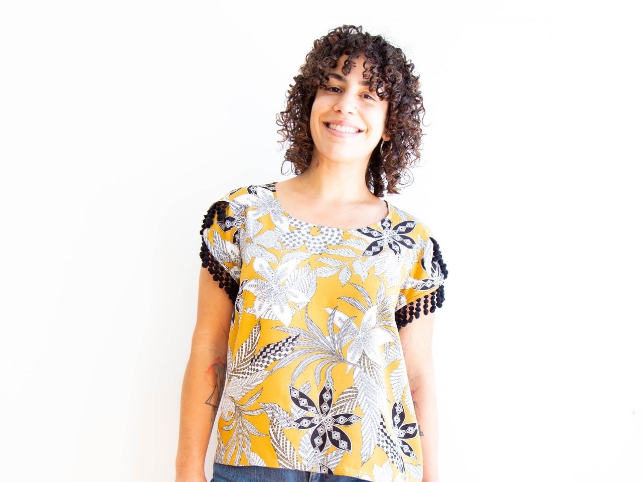 Two views of a Scout Tee with petal sleeves trimmed in black pom-pom fringe, sewn in yellow and white tropical print fabric.