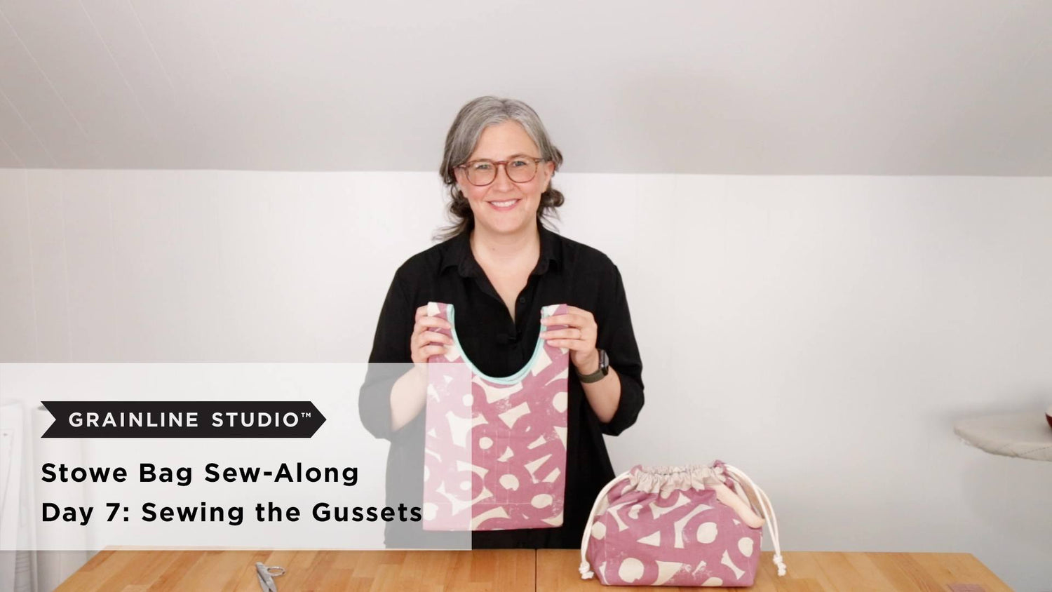 Woman holding up a finished Stowe Bag in mauve printed canvas with teal binding beside a completed Field Bag on the table