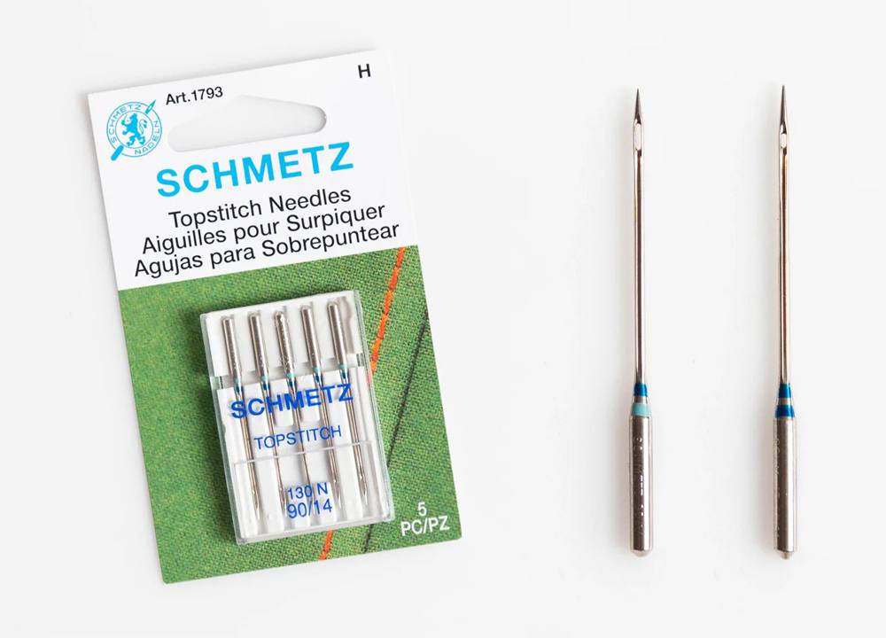 Package of Schmetz topstitch needles shown with two loose needles displaying their large eyes for heavy thread