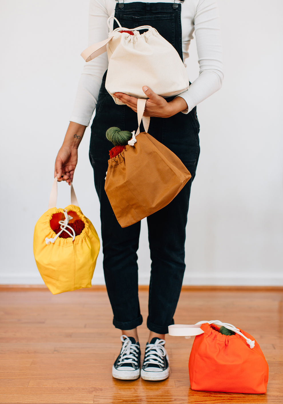 Person holding three canvas field bags in yellow, toffee, and natural, with an orange bag on the floor.