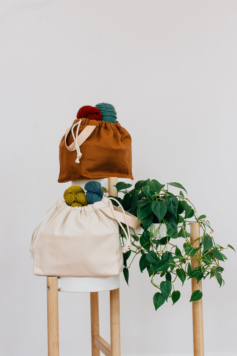 Toffee and natural canvas field bags filled with yarn, staged beside a plant on stools.