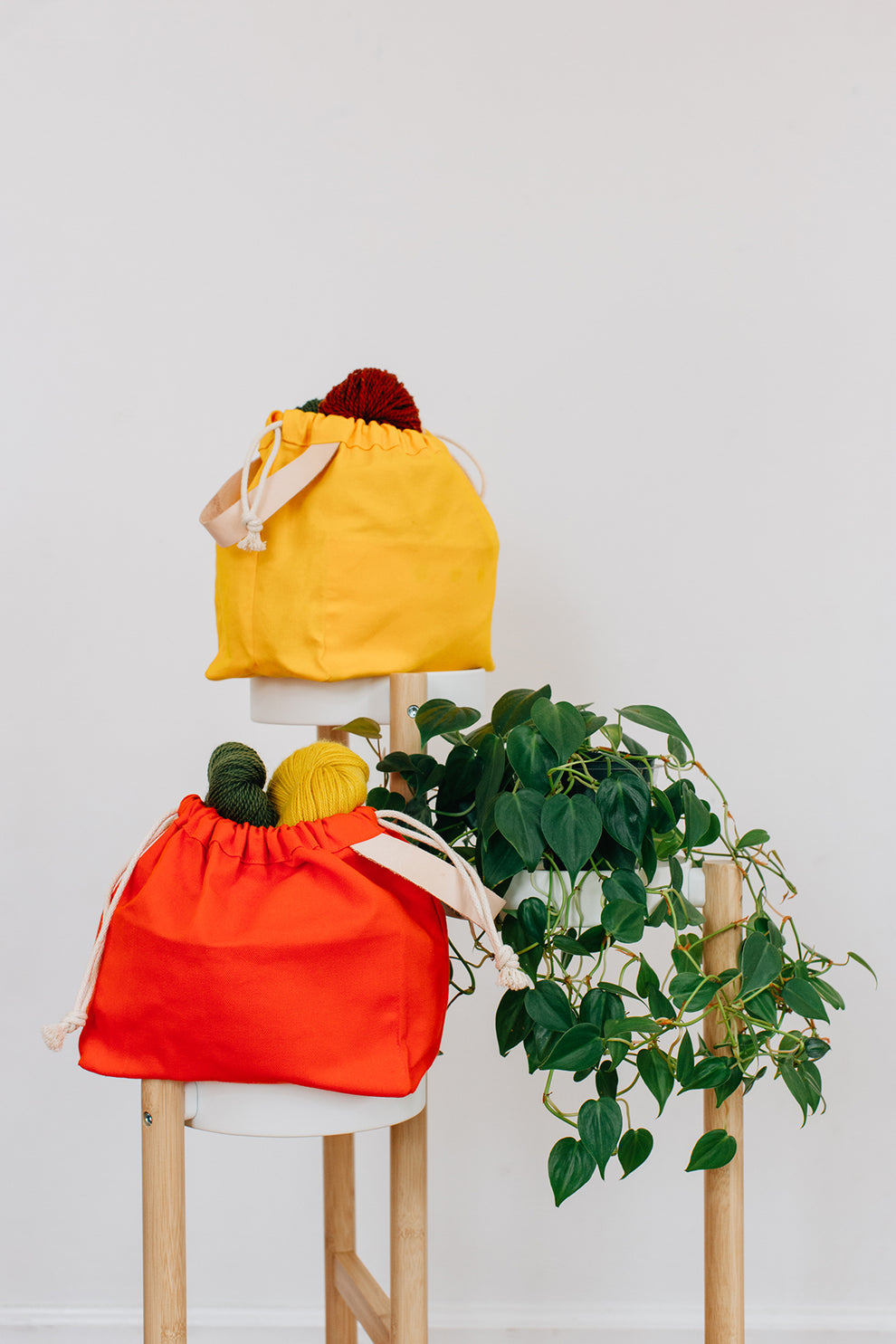 Four field bags in orange, yellow, toffee, and natural cotton filled with yarn, styled with a plant on stools.