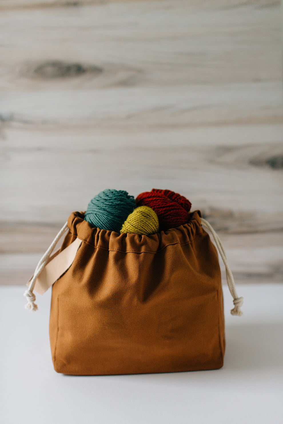 Toffee brown field bag filled with yarn, sitting on a white surface with a wood background.