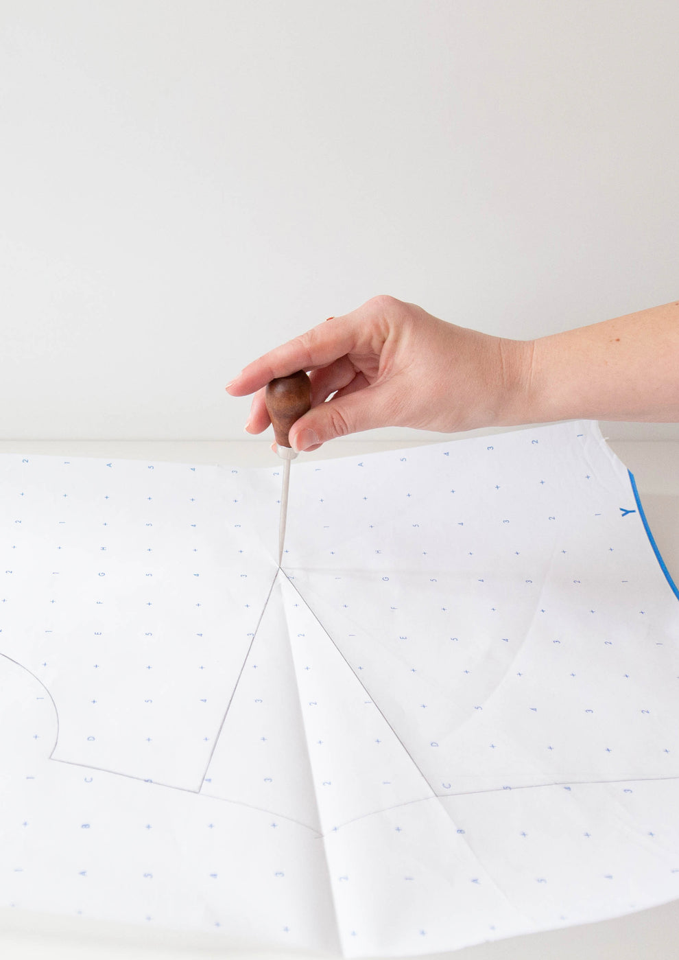 Hand using an awl to mark sewing dart on printed paper pattern with blue grid.