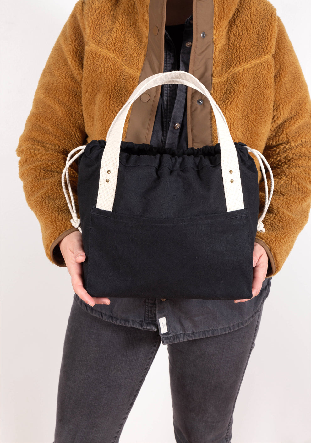 Model wearing tan jacket holding black canvas Town Bag with white handles, front view.