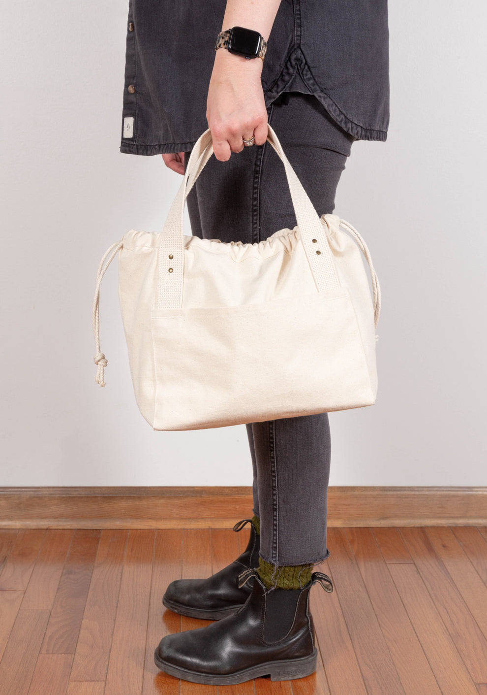 Model standing in black boots holding natural Town Bag at side, drawstring closed.