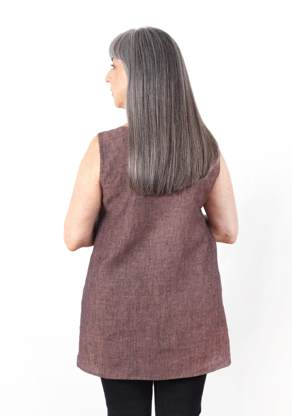 Back view of the Uniform Tunic in sleeveless red-brown linen, modeled with elbows bent.